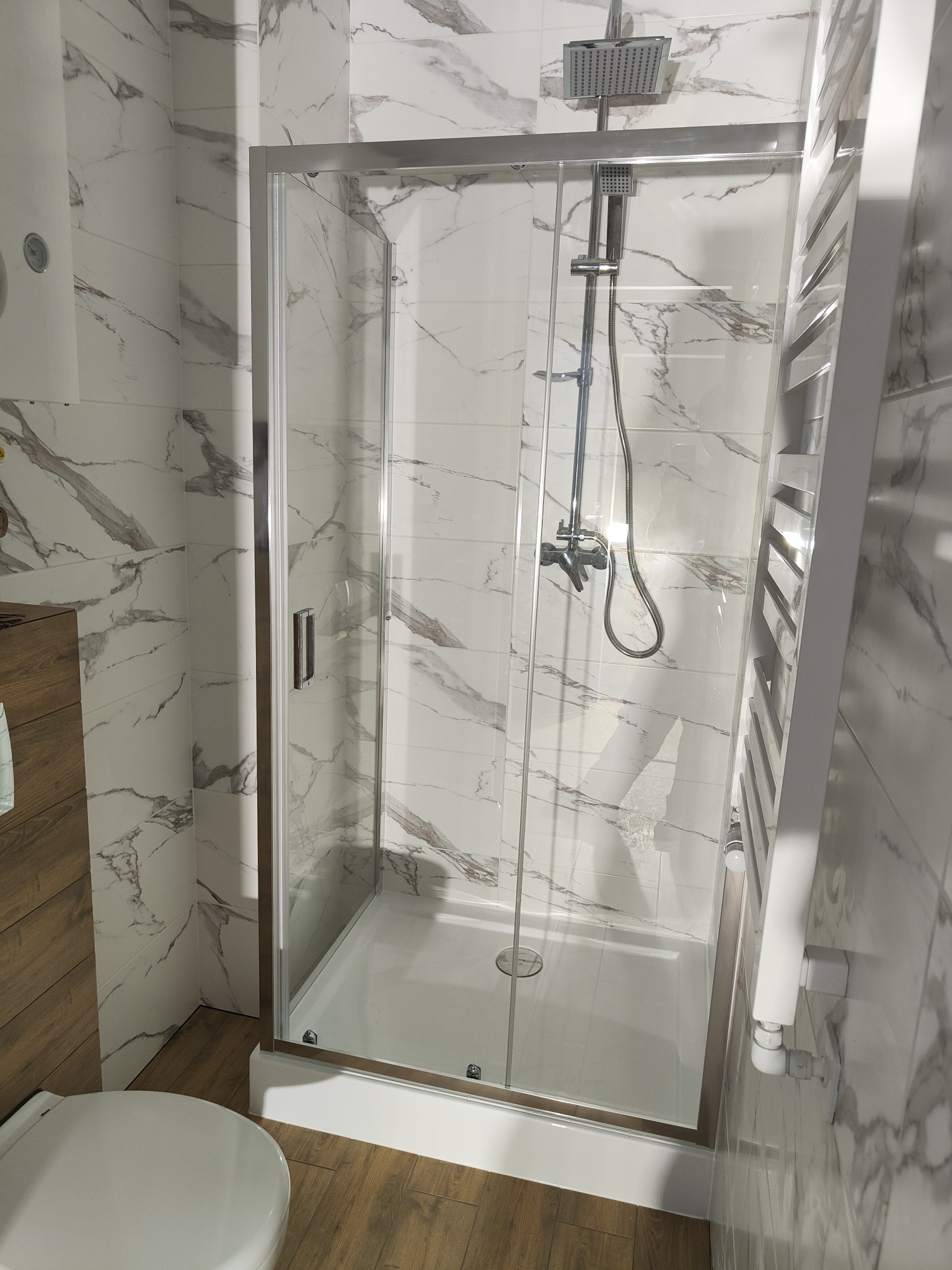 Nowoczesna kabina prysznicowa z chromowanymi detalami i marmurowymi płytkami, zamontowana w jasnej łazience. Widoczny grzejnik drabinkowy.