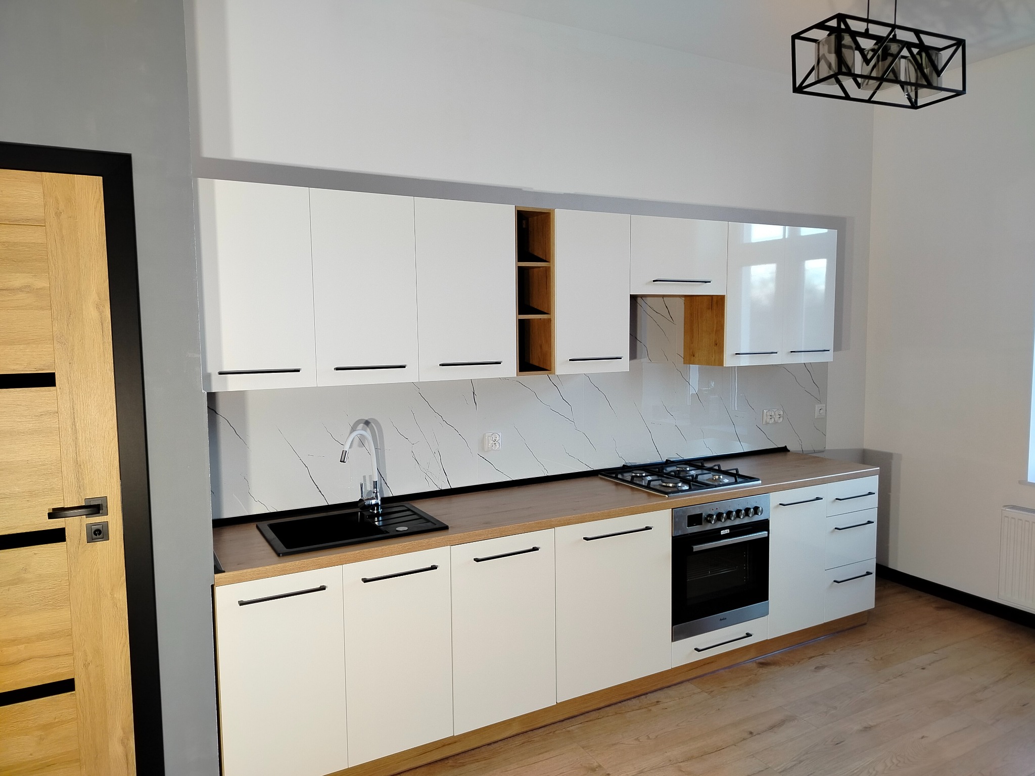 Nowoczesna kuchnia: białe szafki z czarnymi uchwytami, drewniany blat, marmurowe płytki. Czarny zlew i płyta gazowa. Drewniana podłoga i drzwi z czarną ramą.