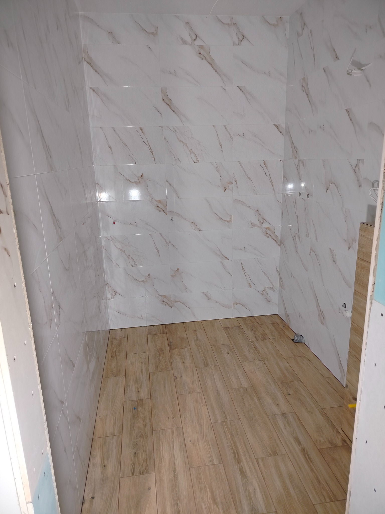 Wykończona łazienka z białymi płytkami imitującymi marmur na ścianach i drewnopodobną podłogą. Widoczne fugi i detale wykończenia. Częściowe ujęcie wnętrza.