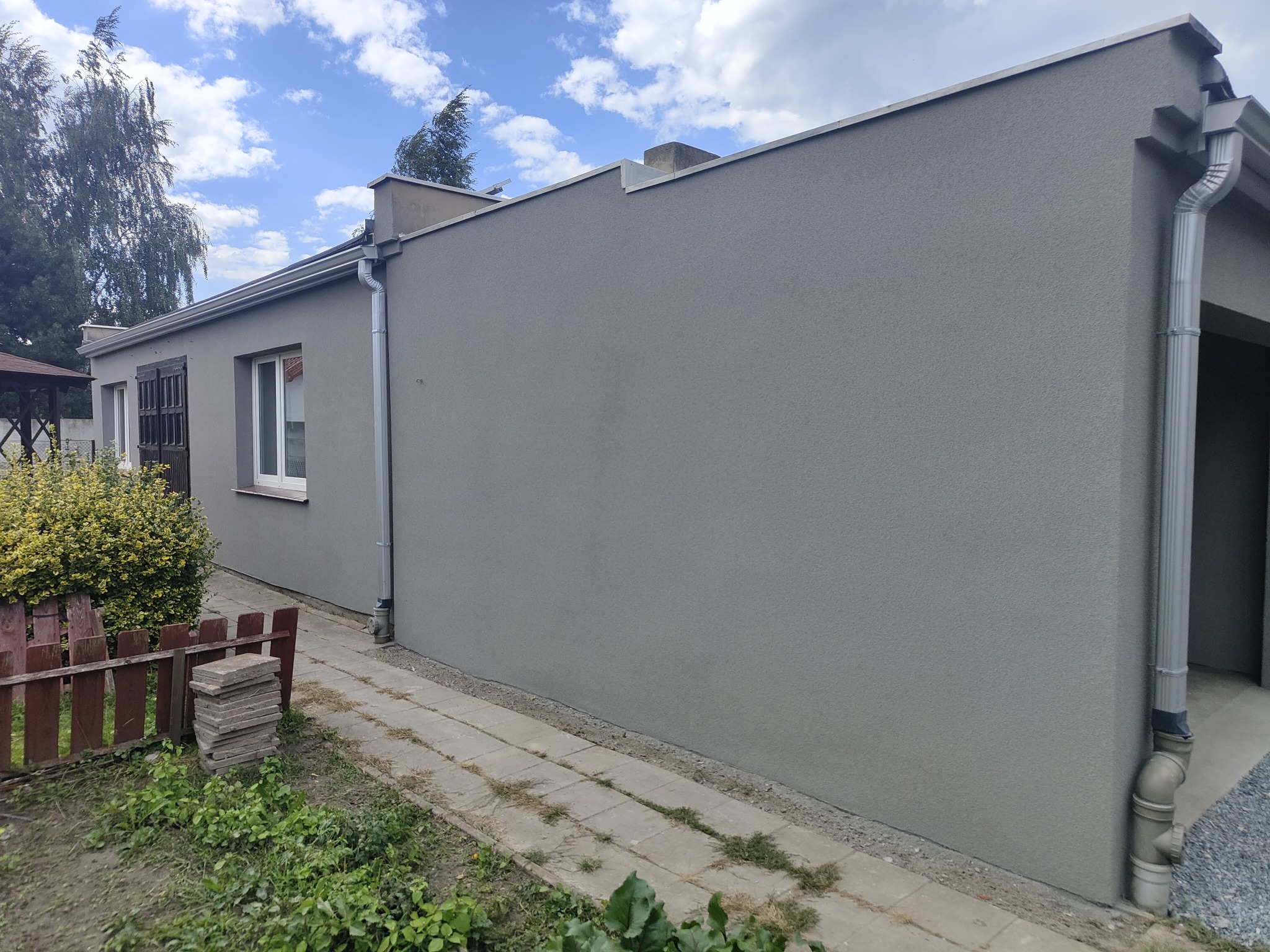 Ściana budynku po tynkowaniu elewacji w szarym kolorze z widoczną rynną, okno z brązowymi okiennicami i fragmentem chodnika z kostki. Budynek jednokondygnacyjny.