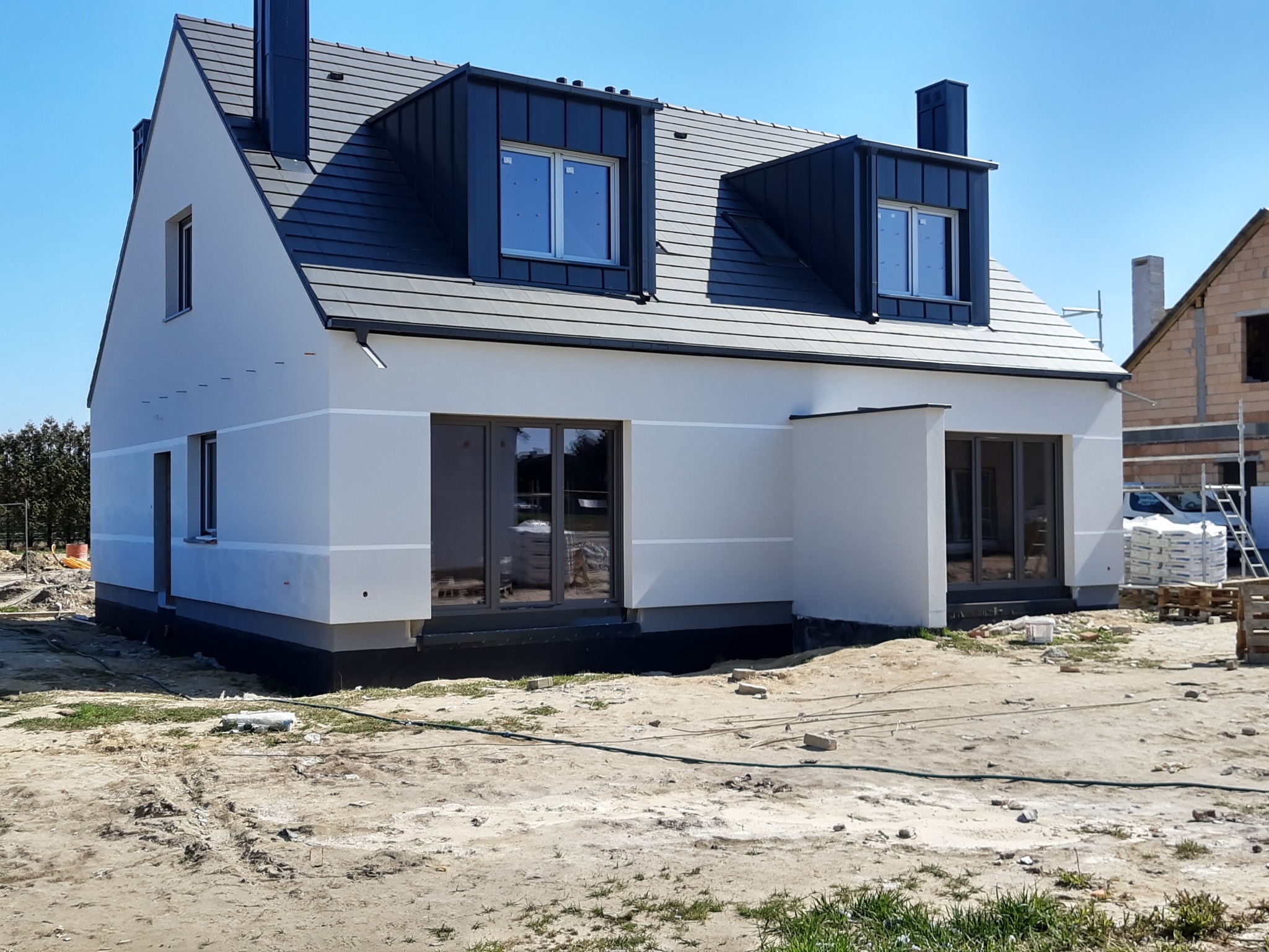 Nowoczesny dom z szarą dachówką i białymi ścianami, widoczny z zewnątrz. Budynek z oknami i kominami, otoczony placem budowy. Częściowo ukończona elewacja.