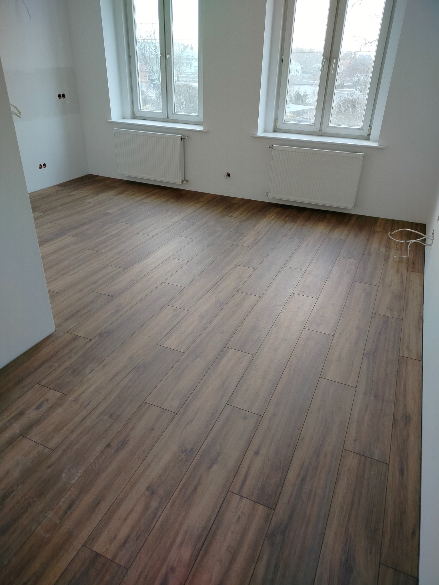 Ułożone panele podłogowe w jasnym pokoju z dwoma oknami. Widoczne grzejniki pod oknami i wystające kable elektryczne ze ściany. Brązowe panele imitujące drewno.