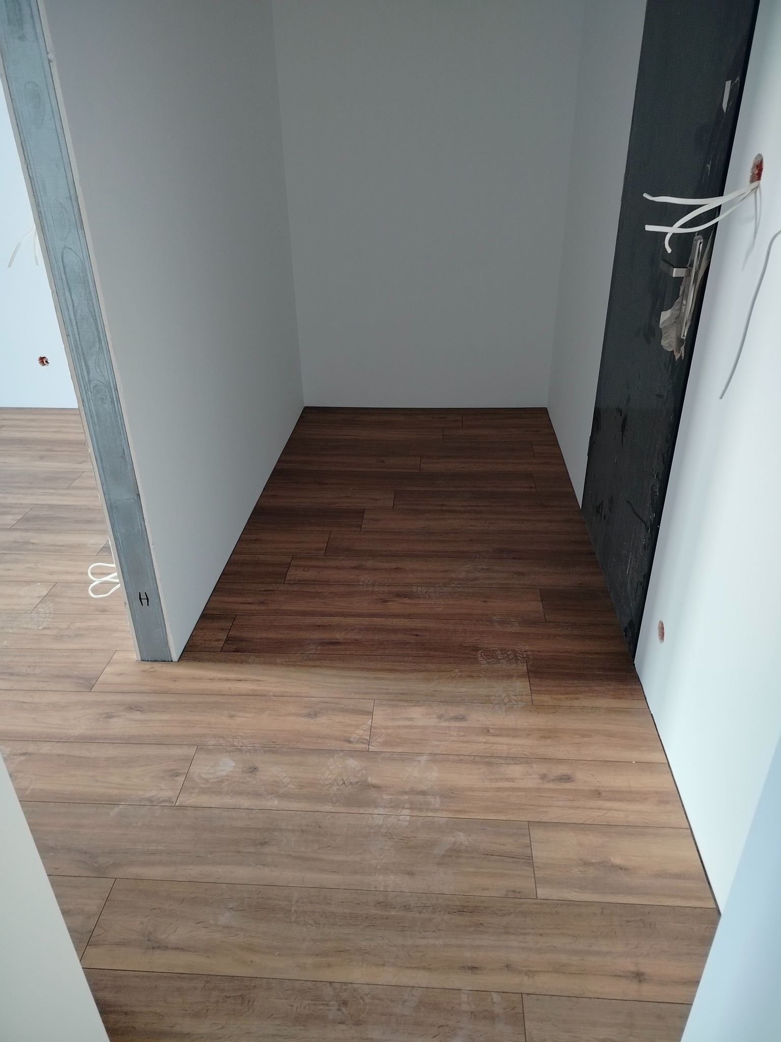 Ukończona podłoga z paneli drewnopodobnych w wąskim pomieszczeniu, widok z góry, fragment metalowej ramy drzwiowej po lewej, instalacja elektryczna po prawej.