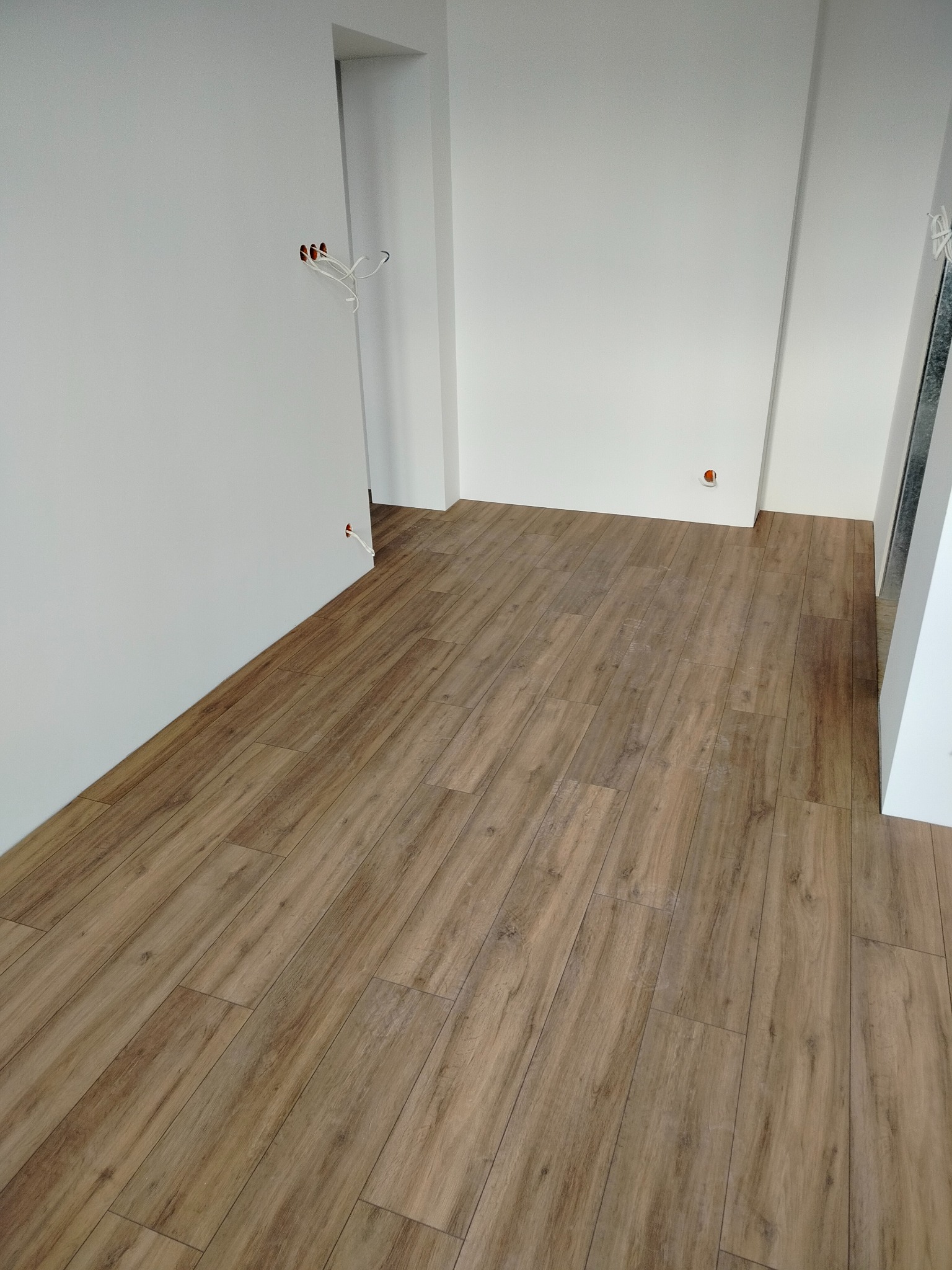 Ułożone panele podłogowe imitujące drewno w jasnym wnętrzu. Widoczne gniazdka elektryczne bez osprzętu na białych ścianach. Kątowe ujęcie.