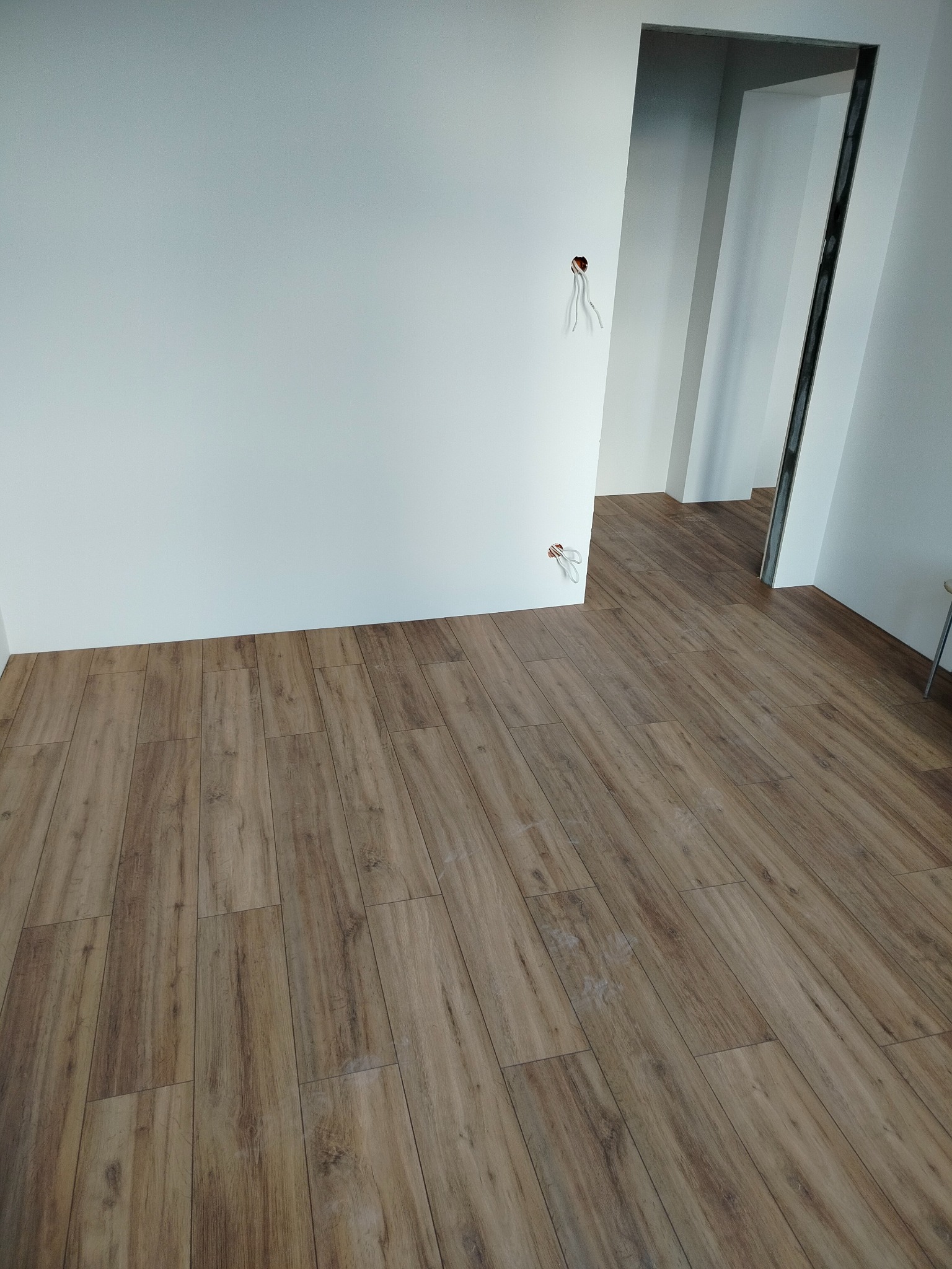 Ułożone panele podłogowe o wzorze drewna w jasnym pomieszczeniu z białymi ścianami. Widoczne wystające kable elektryczne i ościeżnica drzwiowa.