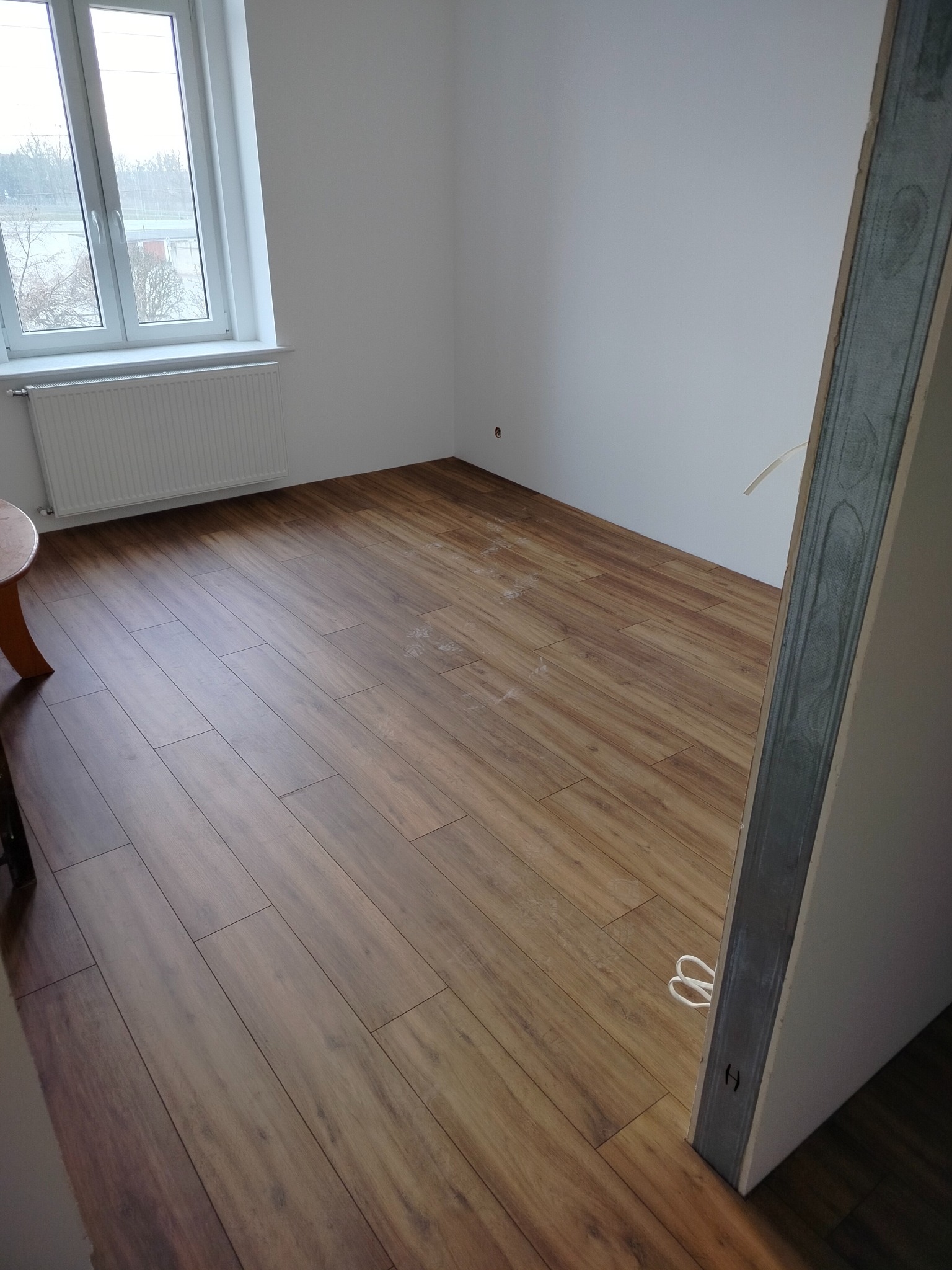 Ukończona podłoga z paneli drewnopodobnych w pokoju z oknem i grzejnikiem. Widoczny fragment futryny drzwi. Naturalne oświetlenie dzienne.