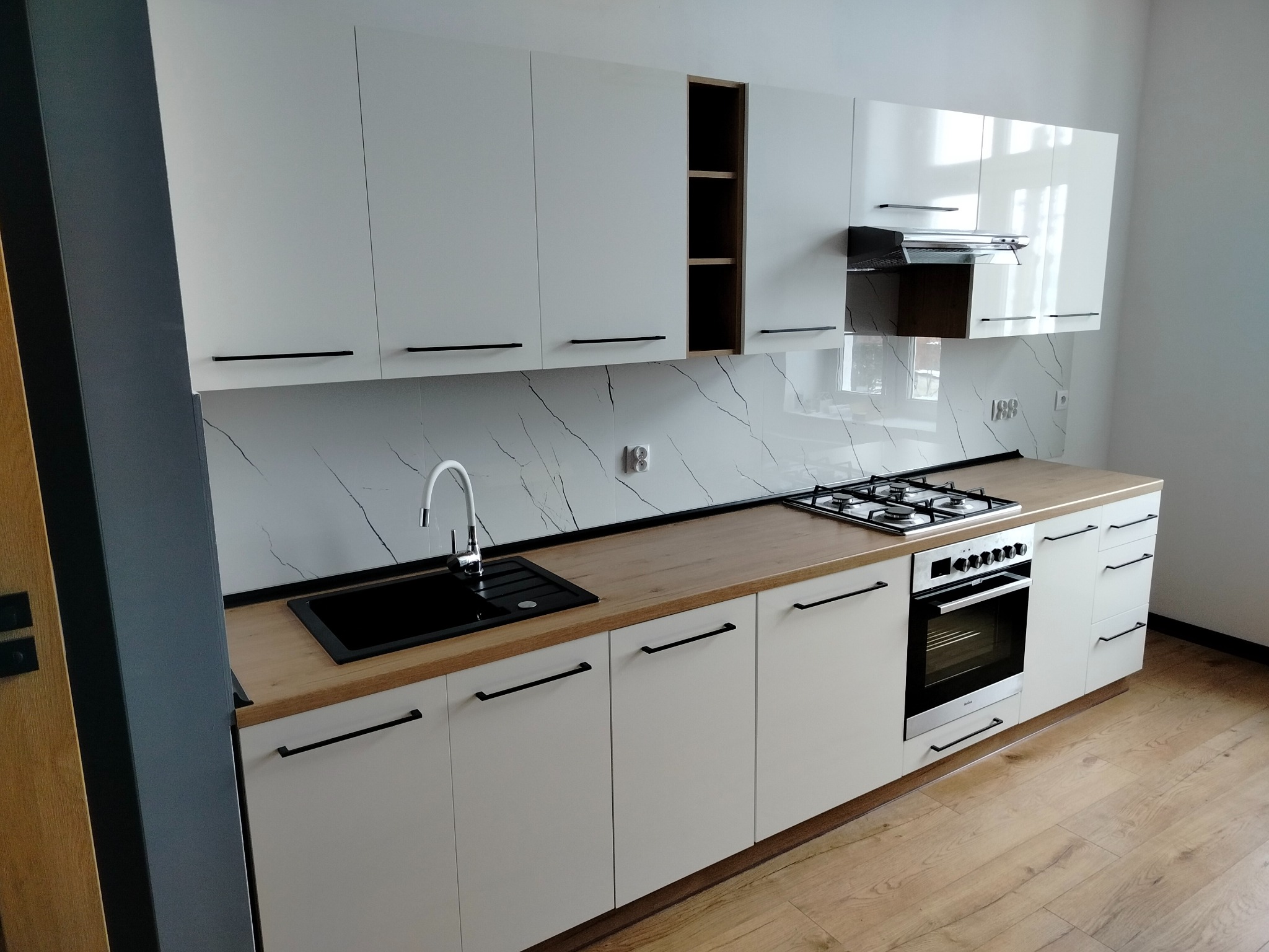 Nowoczesna, jasna kuchnia z białymi szafkami i drewnianym blatem. Czarny zlew i bateria, płyta gazowa z piekarnikiem. Marmurowy backsplash, drewniana podłoga.