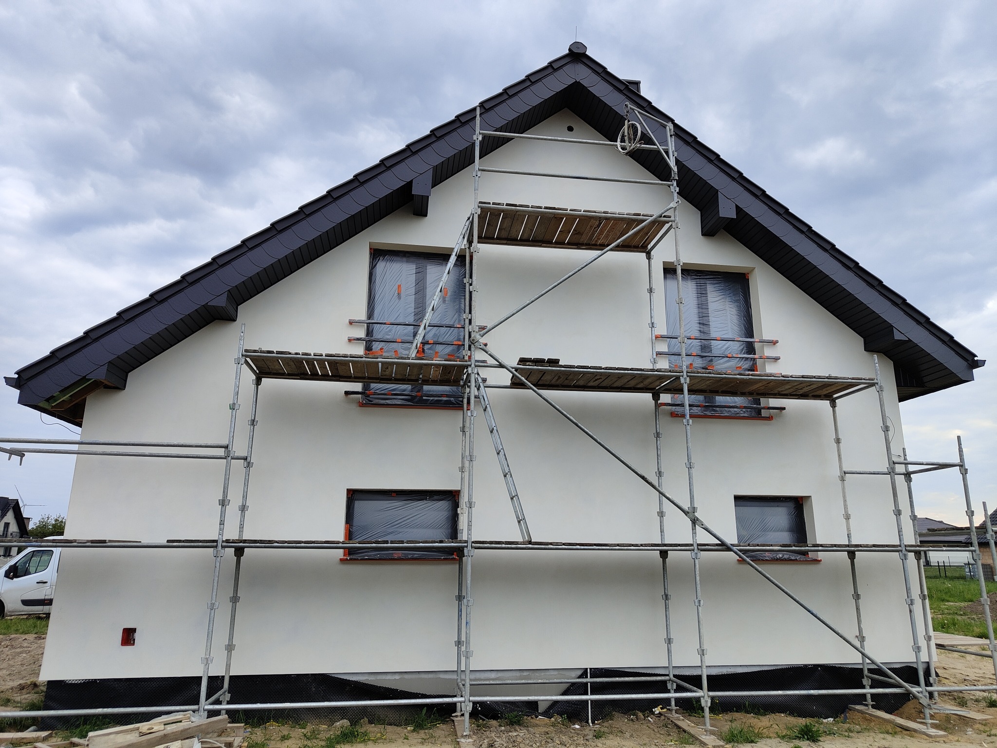 Dom jednorodzinny w trakcie prac elewacyjnych, z oknami zabezpieczonymi folią i rusztowaniem na tle pochmurnego nieba. Widoczne świeżo otynkowane ściany.