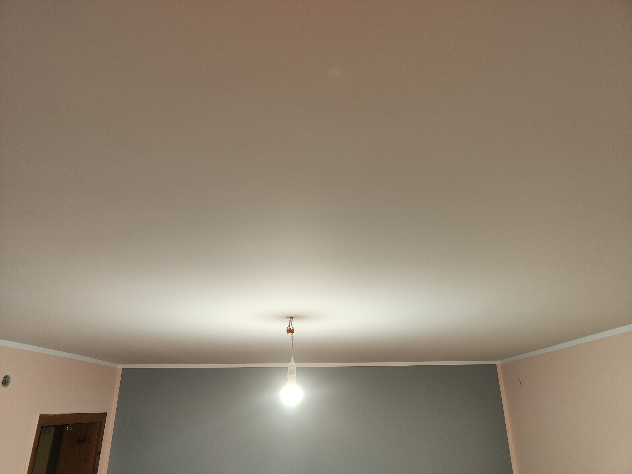 Wnętrze po malowaniu ścian na szaro i różowo, widoczny sufit z zamontowaną żarówką. Biały pasek dekoracyjny na styku ściany i sufitu. Drzwi w lewym dolnym rogu.