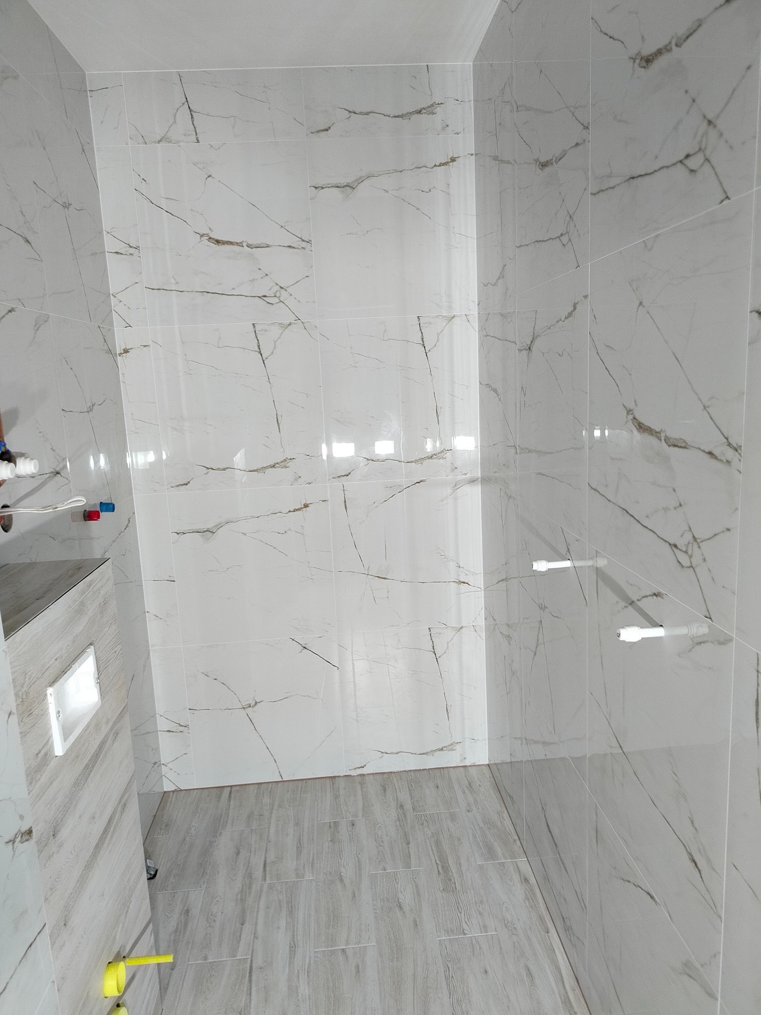 Wykończona łazienka z marmurowymi płytkami na ścianach i podłodze. Widoczne białe rury i elementy instalacji. Minimalistyczny styl, jasne oświetlenie.