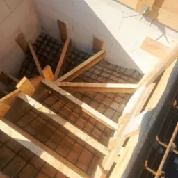 Zbrojenie schodów z drewnianym szalunkiem wewnątrz budynku z białych bloczków. Widoczne stalowe pręty i deski tworzące konstrukcję. Jasne, naturalne oświetlenie.