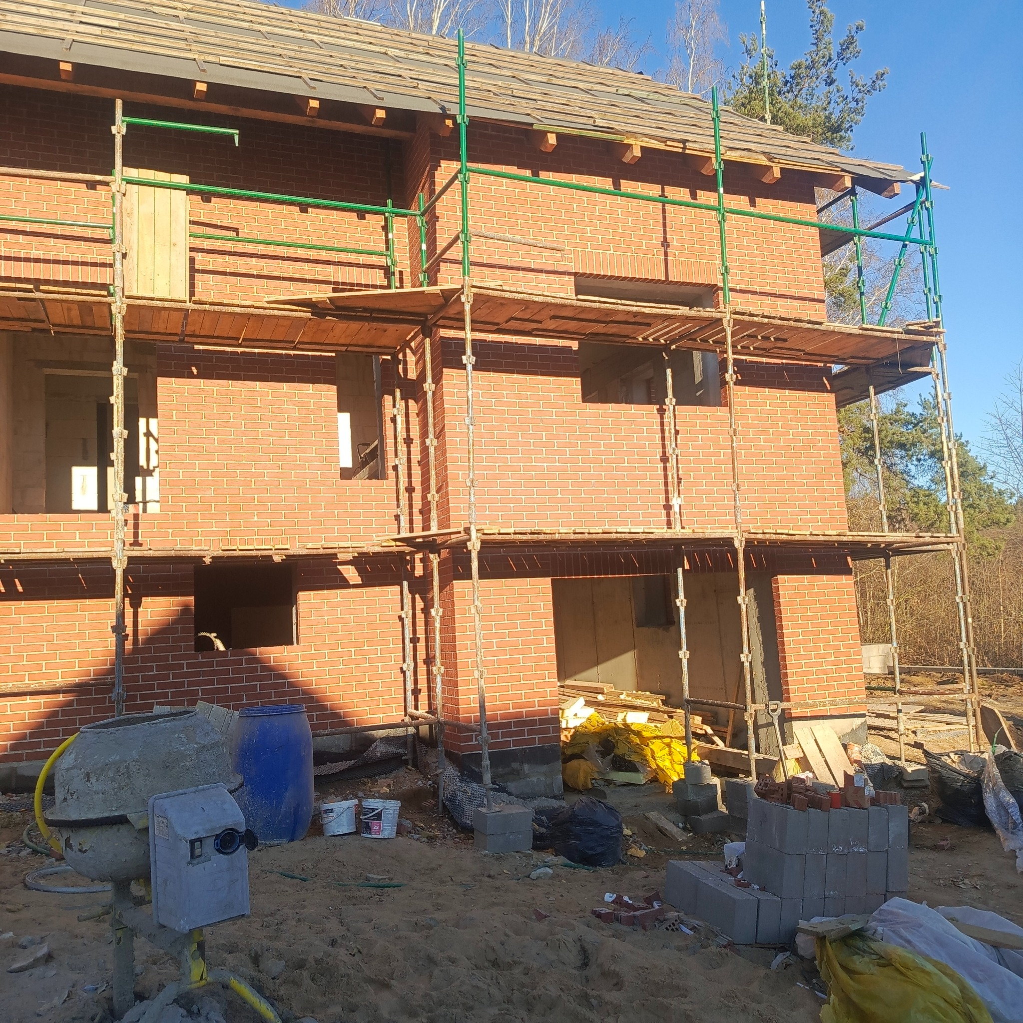 Dwukondygnacyjny dom z czerwonej cegły w trakcie budowy, otoczony rusztowaniami. Widoczna betoniarka i materiały budowlane. Słoneczny dzień.