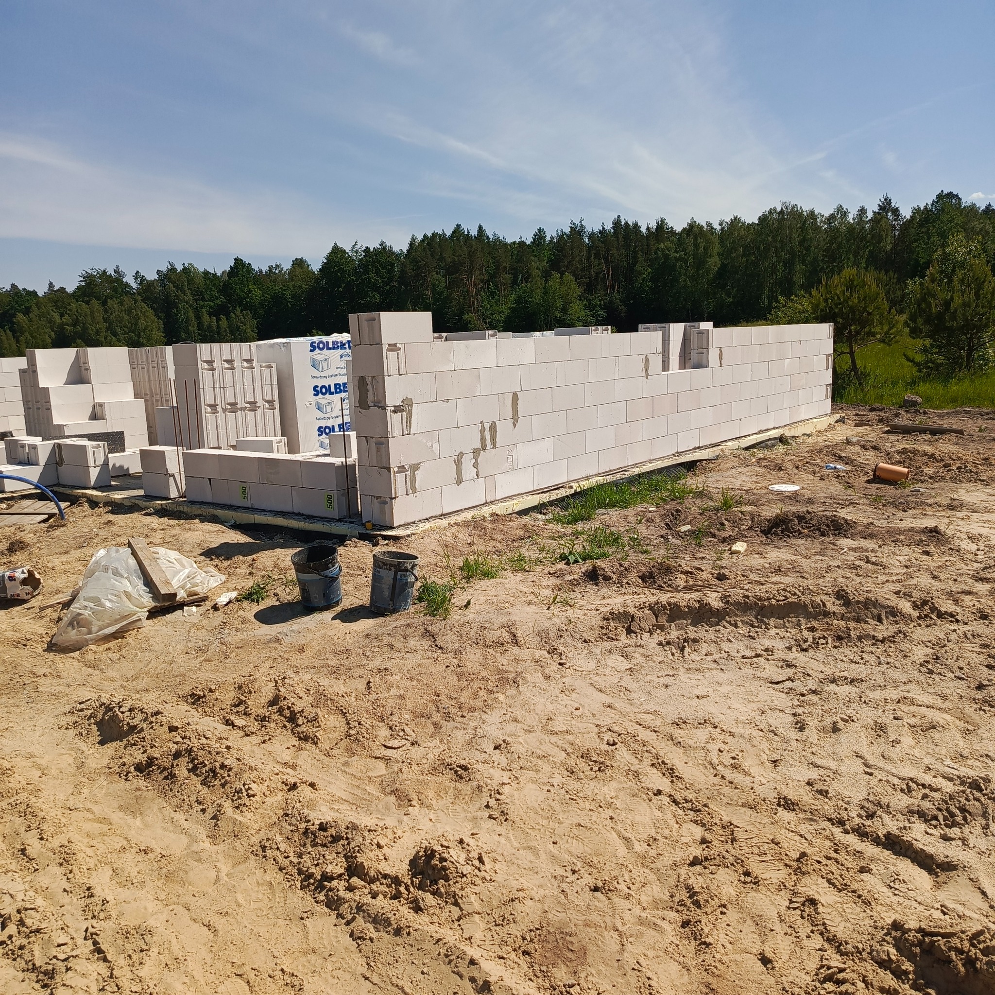 Wczesny etap budowy: wzniesione ściany z białych bloczków Solbet na fundamencie, z widocznymi paletami i narzędziami na piaszczystym podłożu w otoczeniu zieleni.