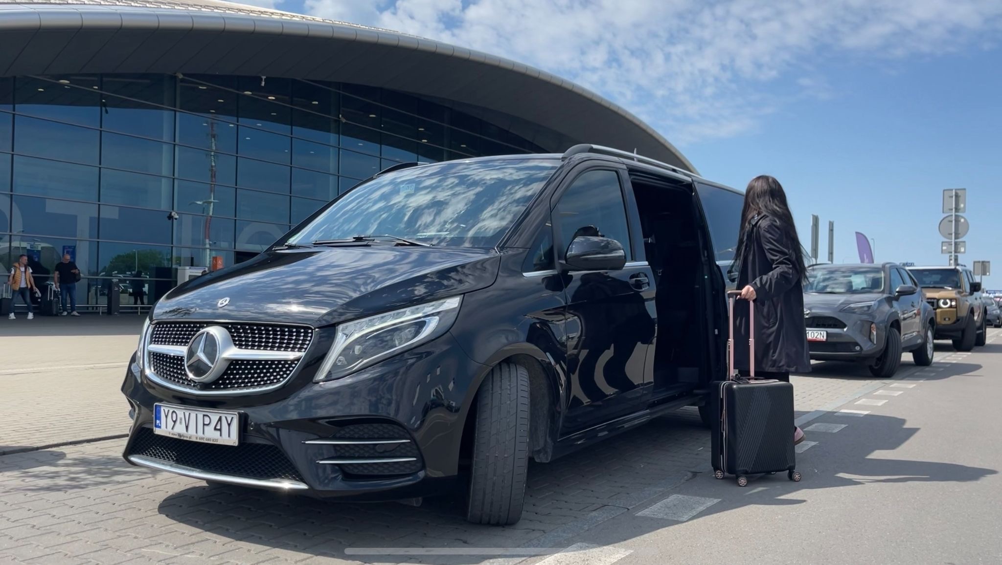 Czarny Mercedes V-klasy z otwartymi drzwiami na lotnisku, kobieta z walizką obok. Tablica rejestracyjna Y9 VIP4Y. W tle budynek lotniska i inne pojazdy.