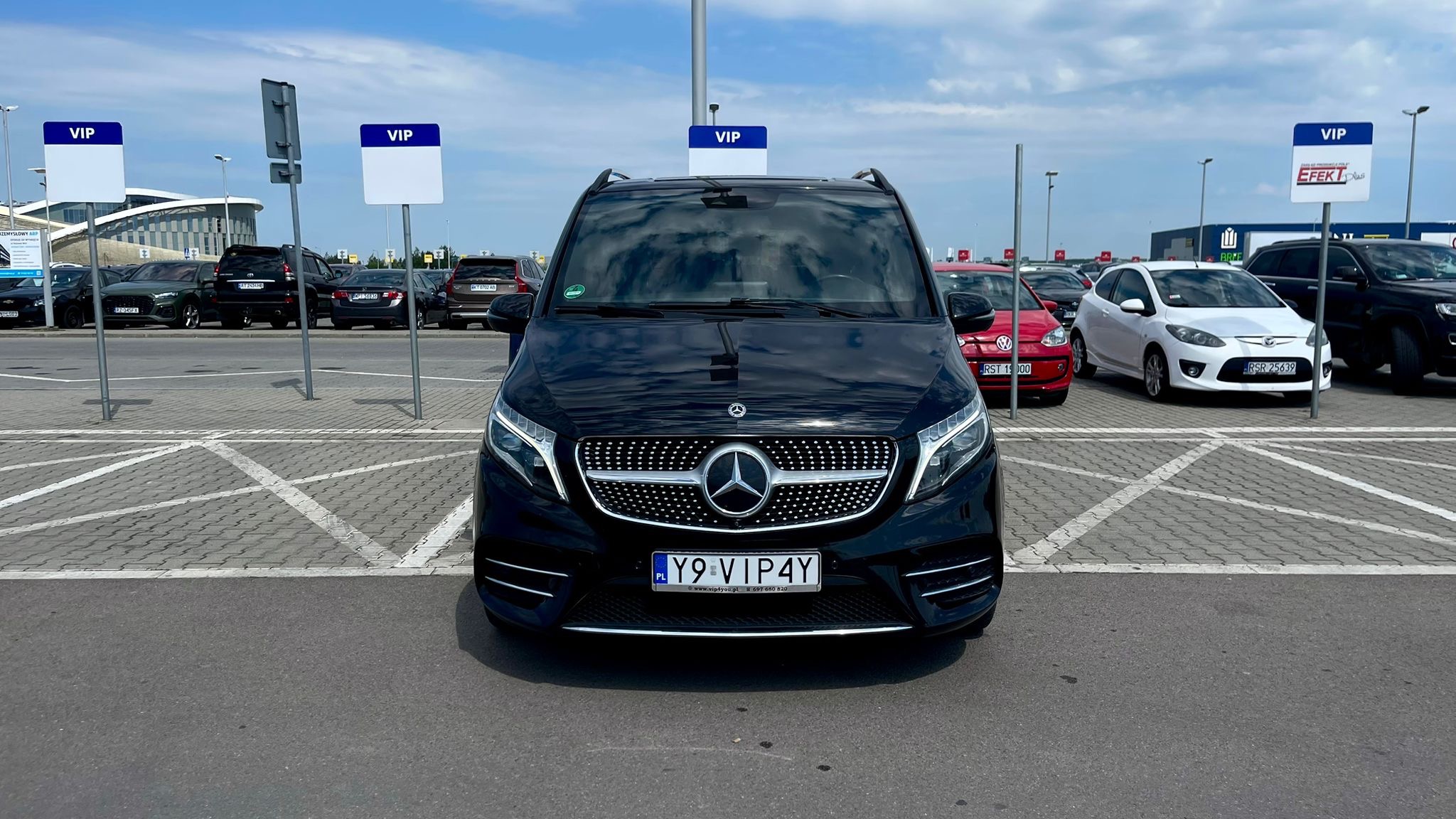 Czarny Mercedes V-klasy z polską rejestracją 'Y9 VIP4Y' na parkingu VIP, z tablicami 'VIP' w tle. Widoczne inne auta i budynek w oddali. Słoneczny dzień.