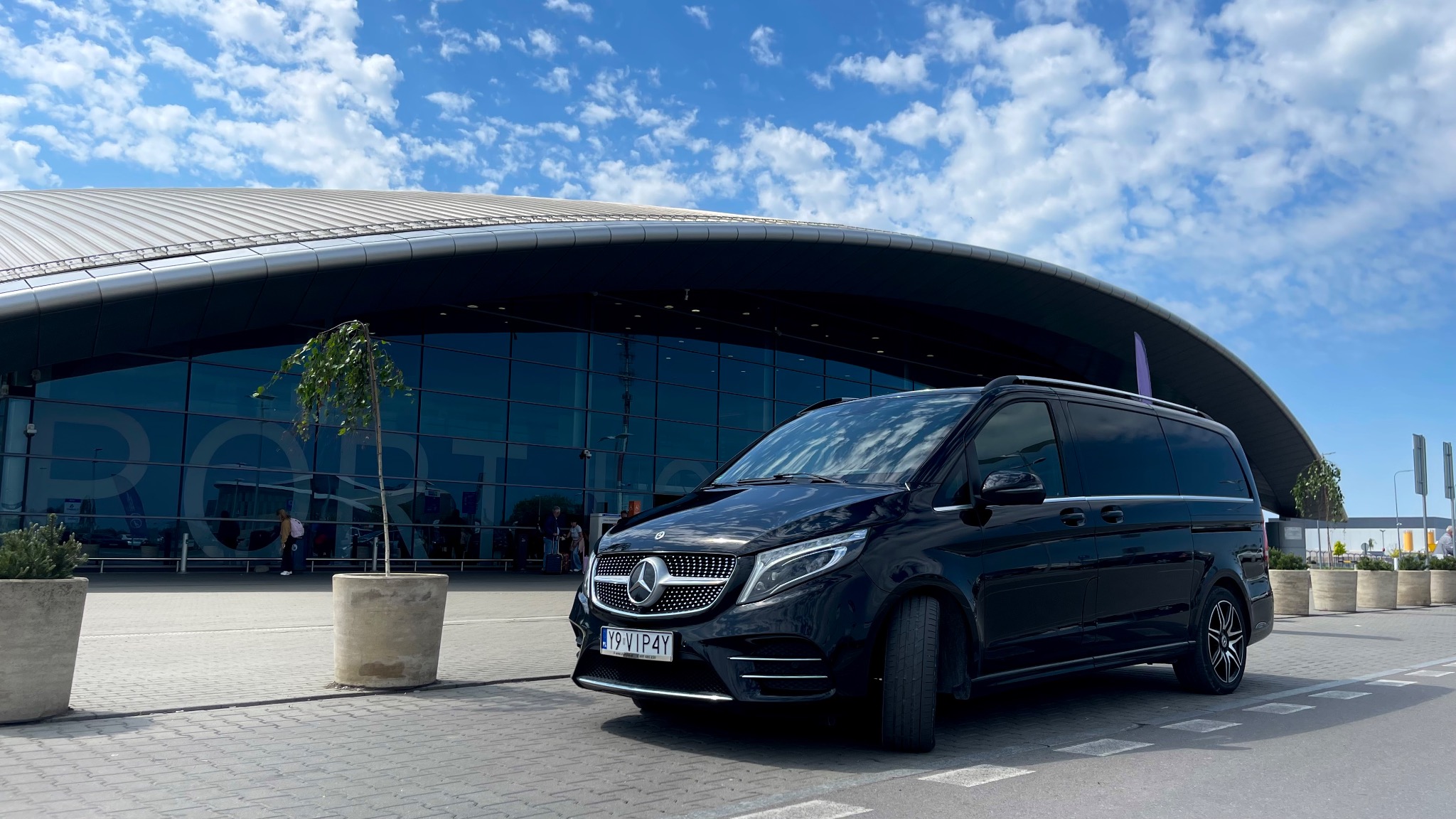 Czarny van Mercedes-Benz V-klasy na tle nowoczesnego budynku lotniska z błękitnym niebem i chmurami. Rejestracja: Y9 VIP4Y. Reprezentacyjny przewóz osób.