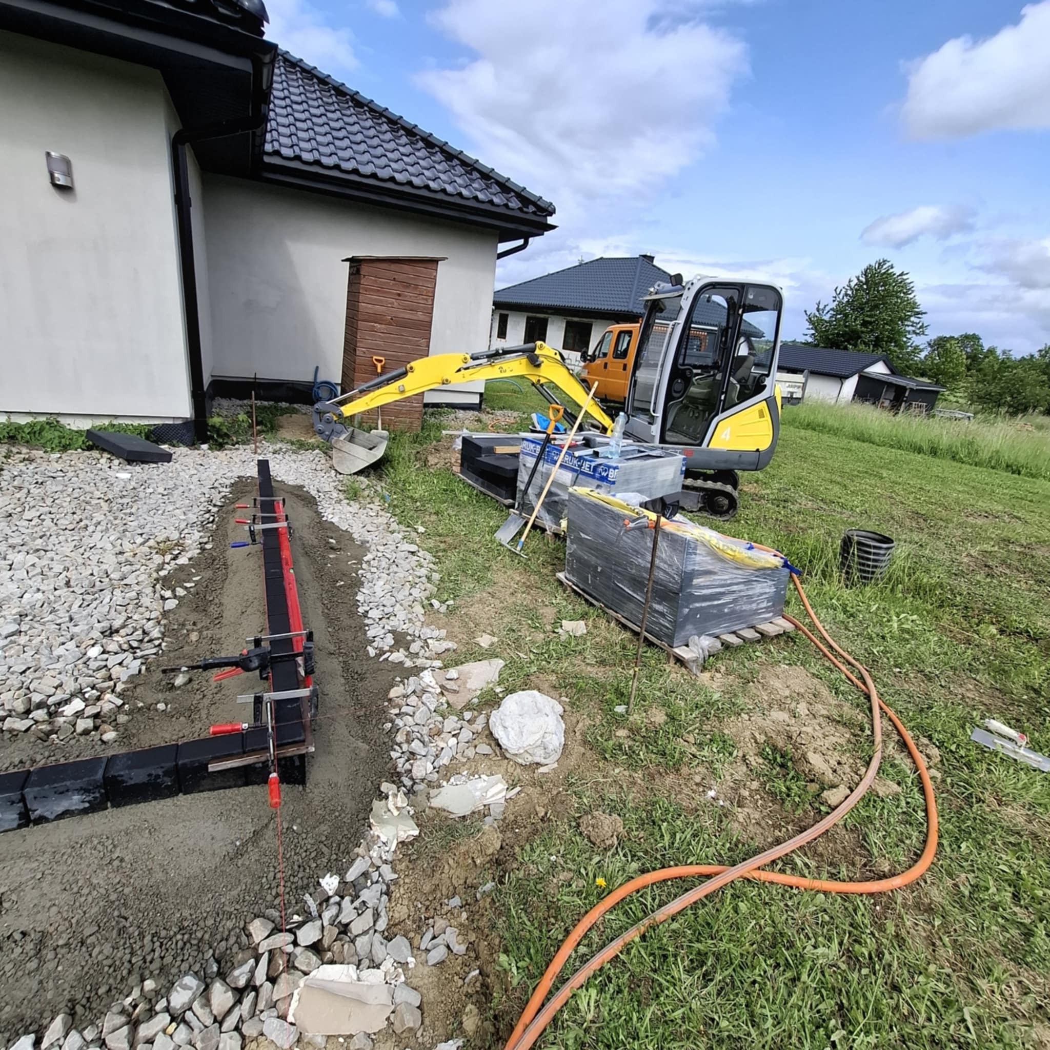 Przygotowanie podłoża pod ścieżkę z kostki: wykop, obrzeża z mocowaniami, mini koparka i paleta z kostką na trawie. Prace ziemne w toku.