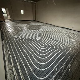PG INSTALACJE PAULINA GAWINOWSKA - Białe rury ogrzewania podłogowego ułożone spiralnie na czarnej siatce w surowym wnętrzu, przygotowane do wylewki. Widoczne kable pod sufitem.