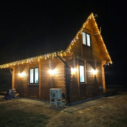 PG INSTALACJE PAULINA GAWINOWSKA - Oświetlony drewniany dom z klimatyzatorem na zewnątrz, nocne ujęcie. Dekoracyjne lampki choinkowe podkreślają kształt dachu.