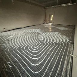 PG INSTALACJE PAULINA GAWINOWSKA - Białe rury ogrzewania podłogowego ułożone spiralnie na czarnej siatce w surowym wnętrzu, widok z góry pod kątem.