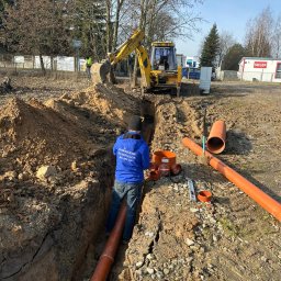 PG INSTALACJE PAULINA GAWINOWSKA - Montaż rur kanalizacyjnych w wykopie ziemnym z użyciem koparki. Widoczny pracownik w niebieskiej bluzie z napisem, pomarańczowe rury i akcesoria.