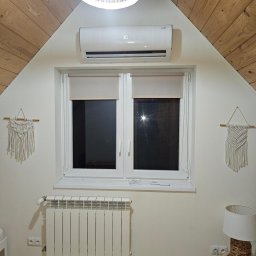 PG INSTALACJE PAULINA GAWINOWSKA - Biały grzejnik aluminiowy pod oknem na poddaszu z drewnianą boazerią, klimatyzator ścienny, lampa i makramy na ścianie.