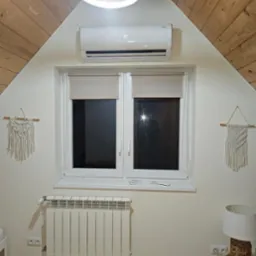Biały grzejnik aluminiowy pod oknem na poddaszu z drewnianą boazerią, klimatyzator ścienny, lampa i makramy na ścianie.