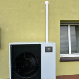 Biała pompa ciepła Mitsubishi Zubadan R32 zamontowana przy żółtej ścianie budynku z oknem. Widoczna biała rura odprowadzająca.