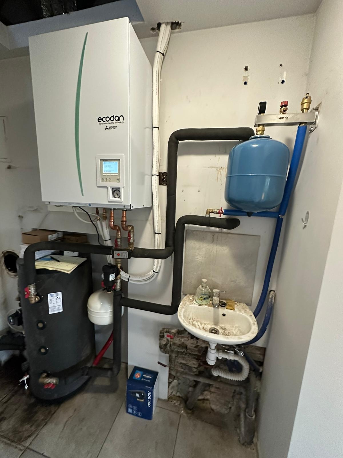 Ecodan Mitsubishi Electric pompa ciepła, zbiornik buforowy i rury w kotłowni. Widoczna umywalka i instalacja hydrauliczna.