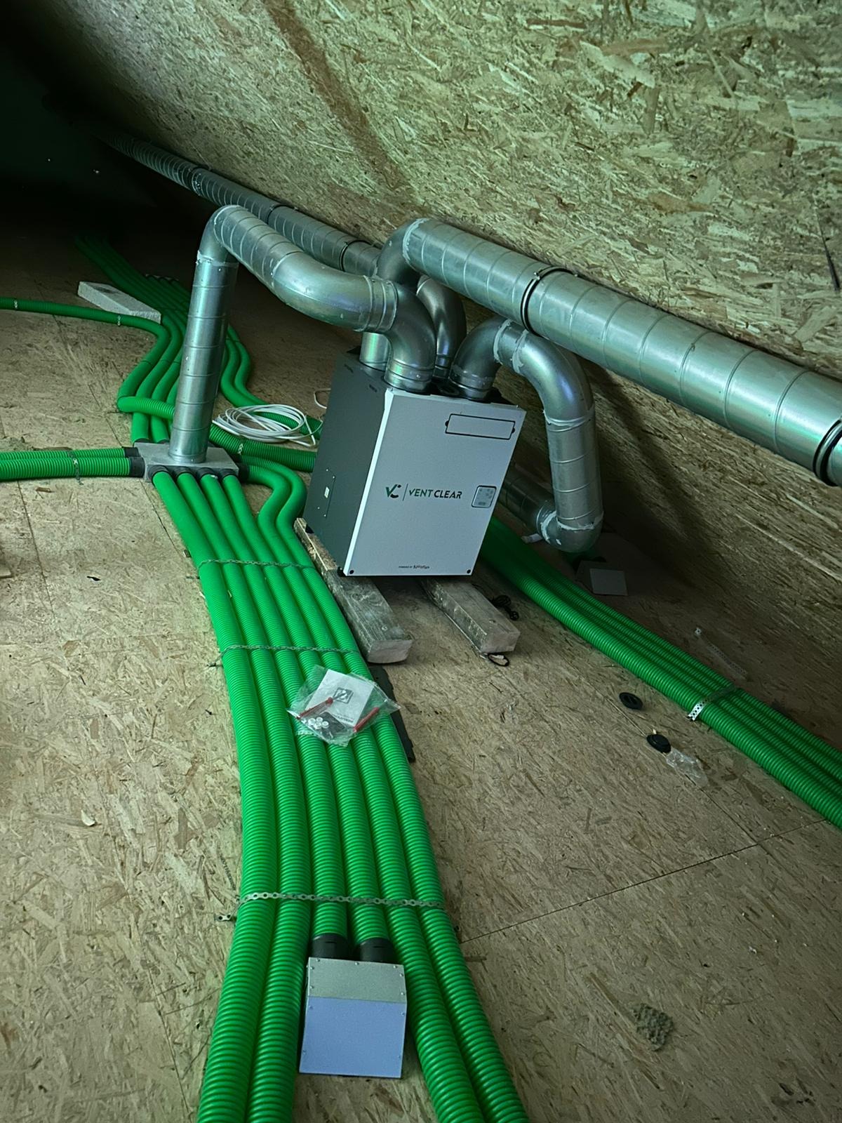 Szara centrala wentylacyjna Vent Clear z podłączonymi, zielonymi rurami wentylacyjnymi na poddaszu z drewnianą boazerią, widoczne srebrne rury.