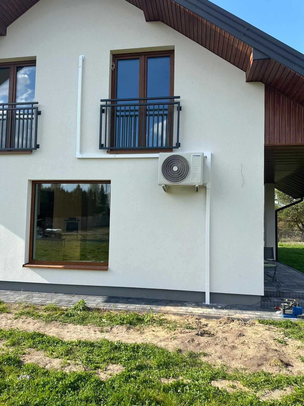 Biała elewacja domu z zamontowanym klimatyzatorem Hyundai, rurami odprowadzającymi skropliny i brązowymi oknami z balkonami. Widok z poziomu gruntu.