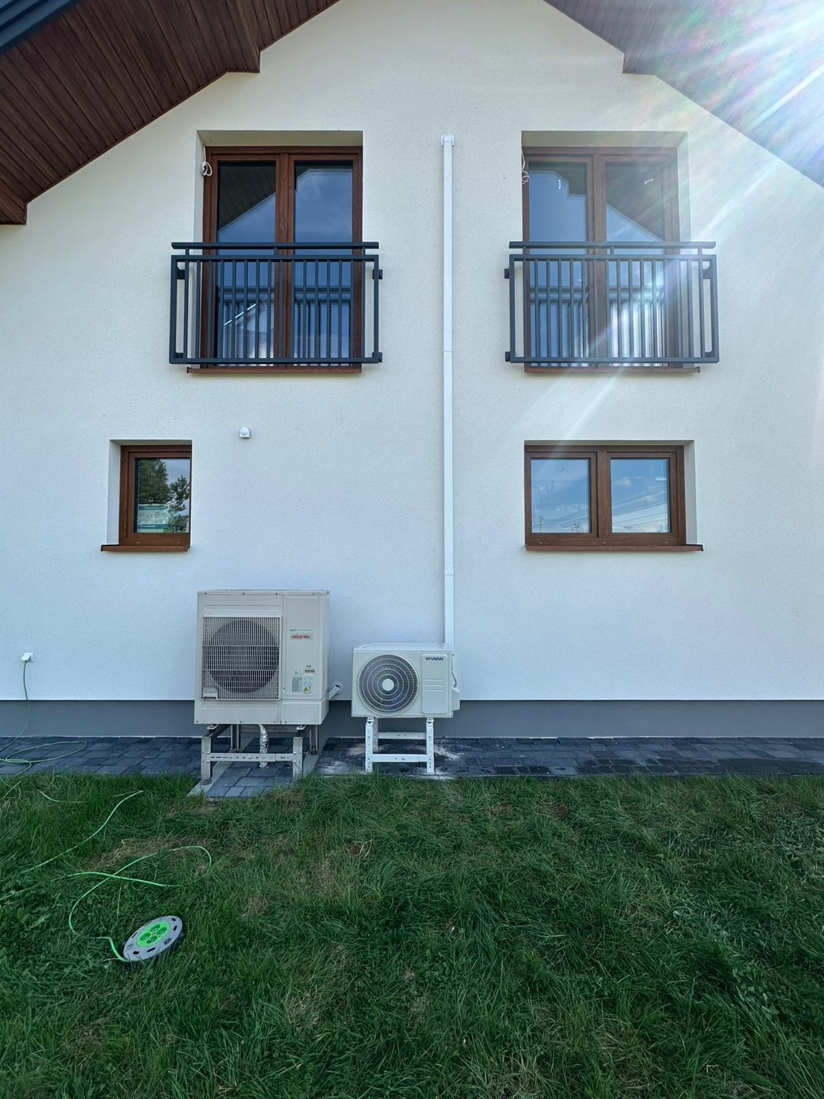 Biały dom z dwoma klimatyzatorami (Atlantic i Hyundai) na zewnątrz. Widoczne okna z brązowymi ramami i czarnymi balkonami. Na trawie leży zielony przedłużacz.