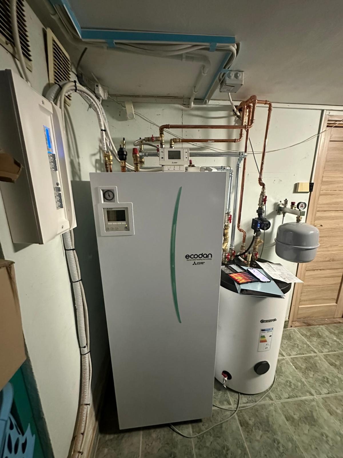 Biały piec Ecodan z zielonym akcentem, obok biały zbiornik Kospel. Widoczne miedziane rury i zawory. Instalacja w pomieszczeniu z zieloną ścianą i płytkami na podłodze.