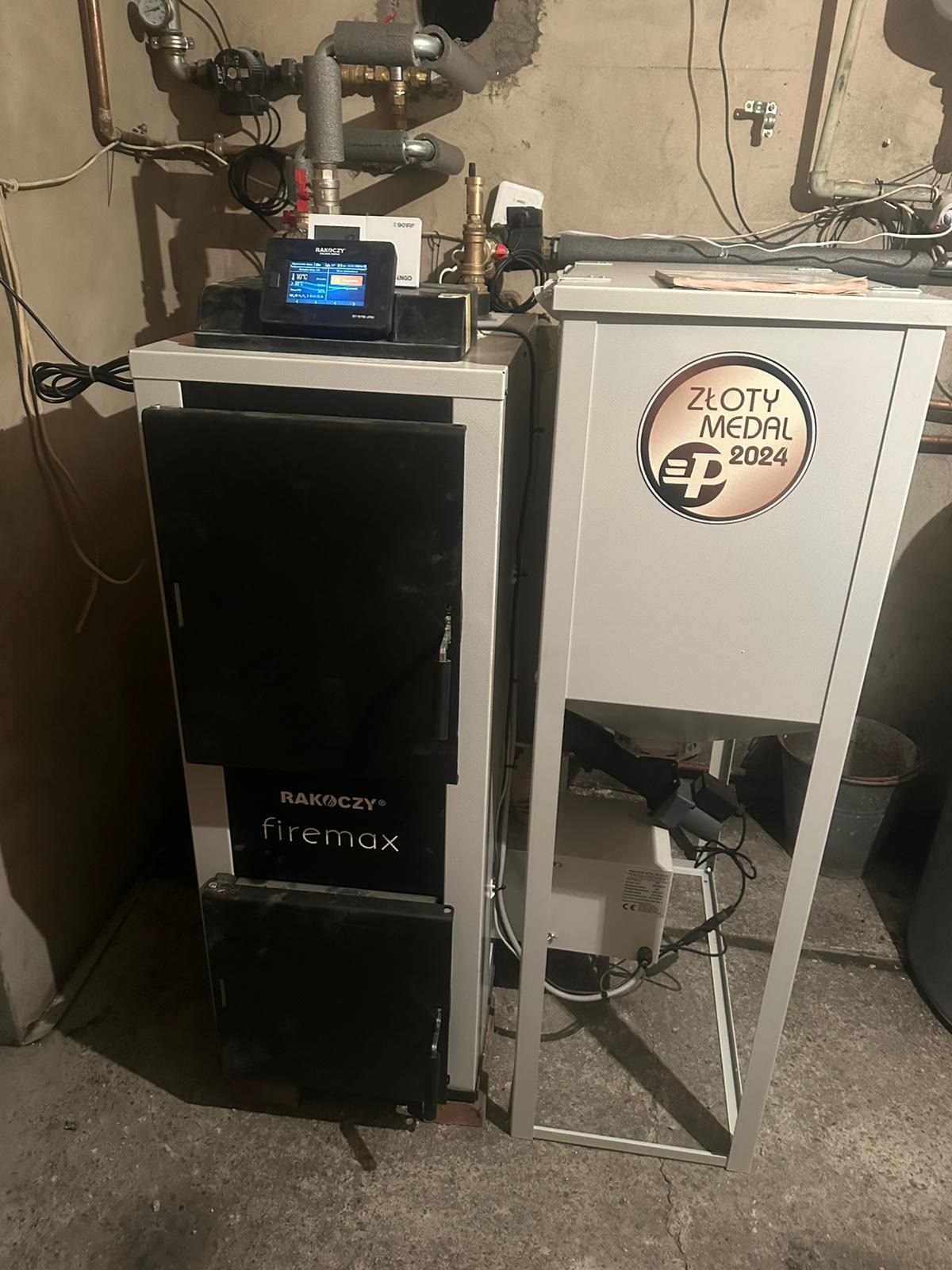 Nowoczesny piec Rakoczy Firemax z podajnikiem, oznaczony 'Złotym Medalem 2024', zainstalowany w kotłowni. Widoczne rury instalacji CO i panel sterowania.