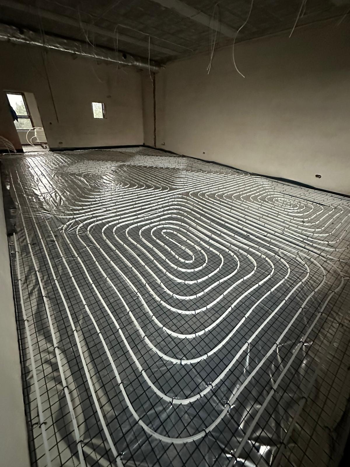 Białe rury ogrzewania podłogowego ułożone spiralnie na czarnej siatce w surowym wnętrzu, przygotowane do wylewki. Widoczne kable pod sufitem.