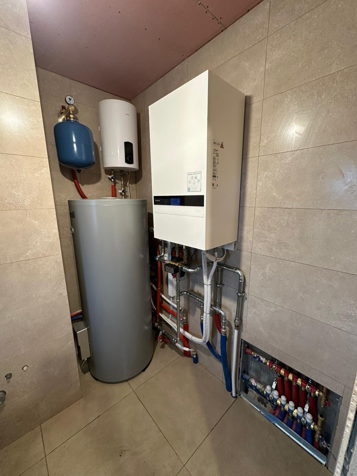 Nowoczesna kotłownia z piecem gazowym, zasobnikiem, naczyniem przeponowym i rozdzielaczem w pomieszczeniu wykończonym płytkami ceramicznymi.