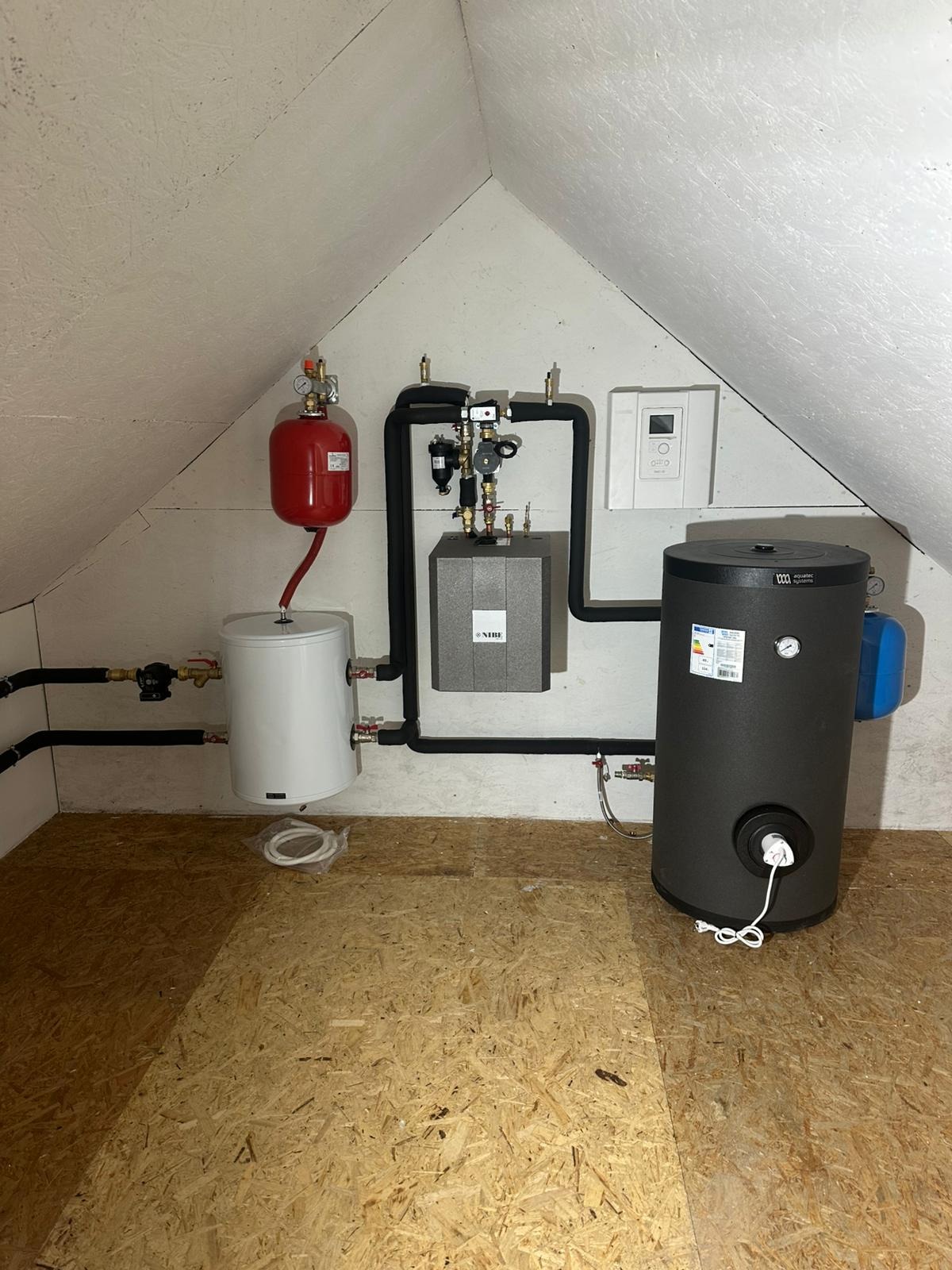 Zestaw instalacji grzewczej w poddaszu: dwa zbiorniki, centrala sterująca NIBE, czarne rury. Całość zamontowana na ścianie pod skosem.