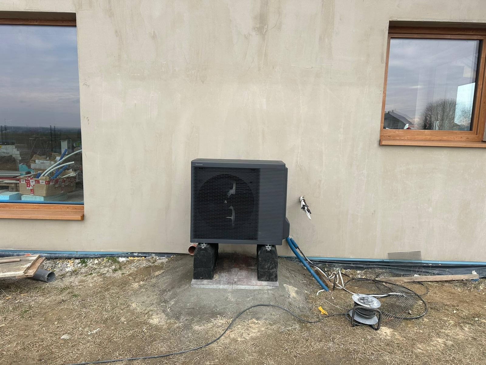 Zamontowana pompa ciepła Panasonic na betonowej podstawie przy ścianie budynku z dwoma oknami. Widoczne przewody i elementy instalacji.