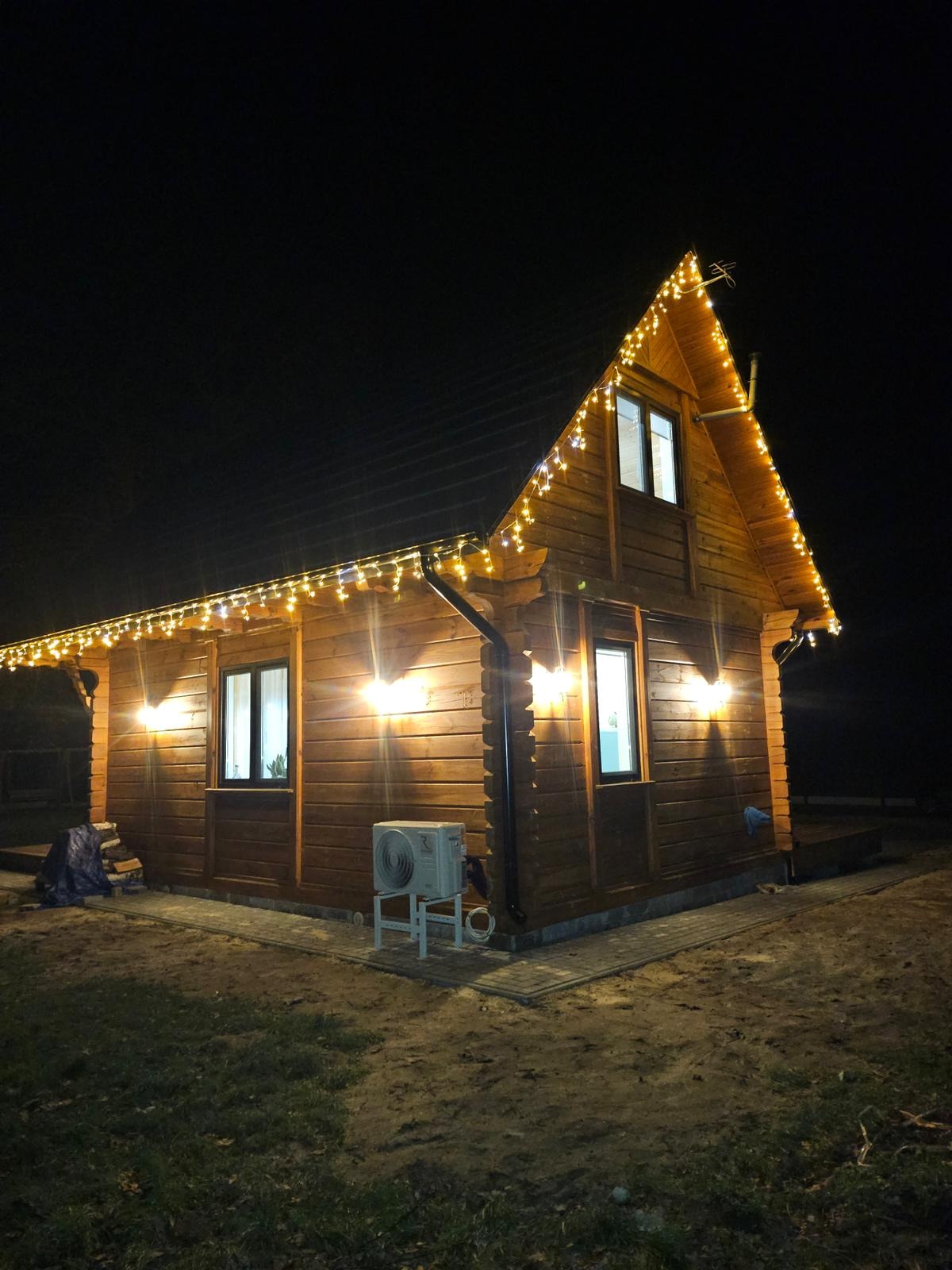 Oświetlony drewniany dom z klimatyzatorem na zewnątrz, nocne ujęcie. Dekoracyjne lampki choinkowe podkreślają kształt dachu.