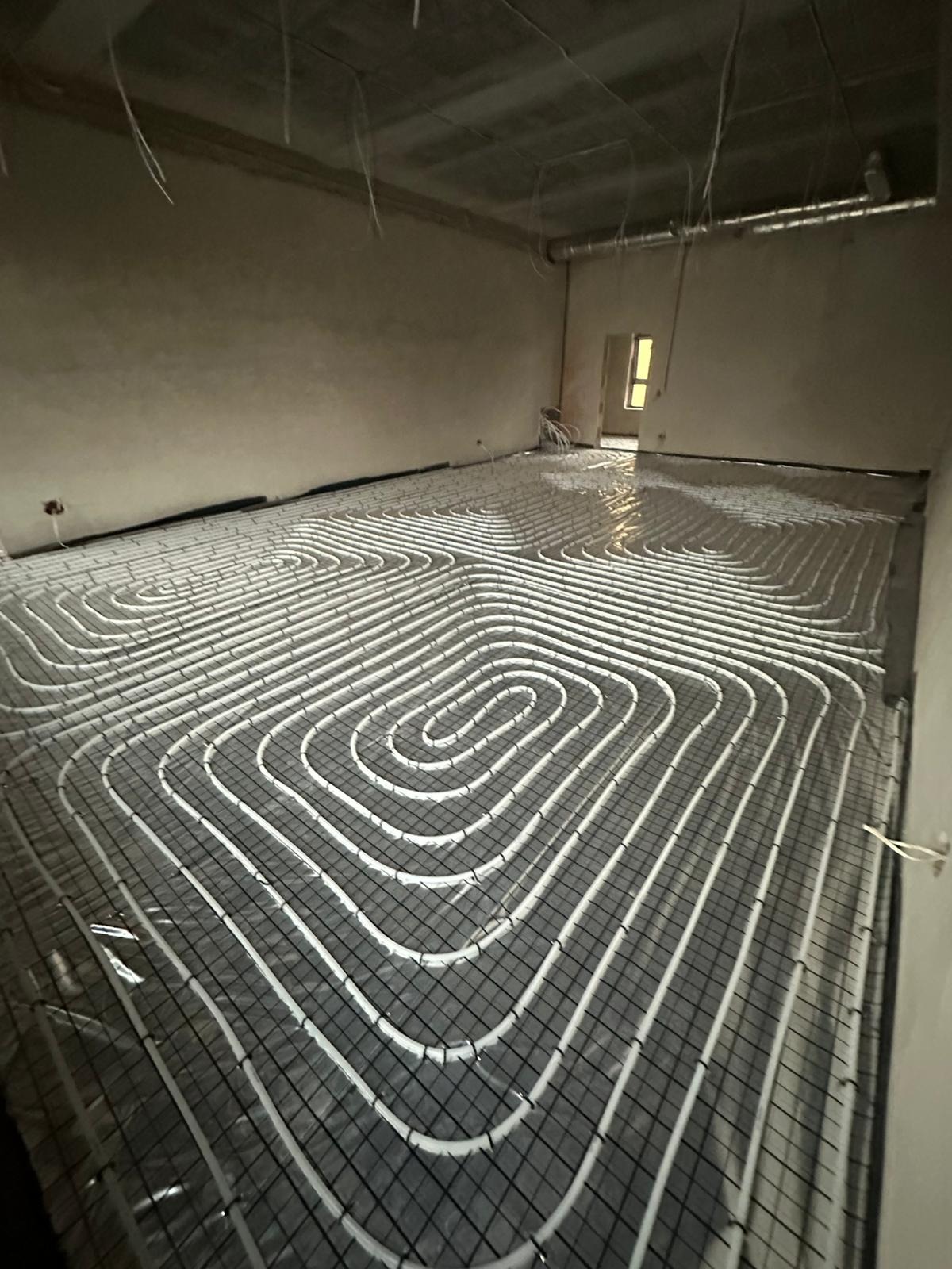 Białe rury ogrzewania podłogowego ułożone spiralnie na czarnej siatce w surowym wnętrzu, widok z góry pod kątem.