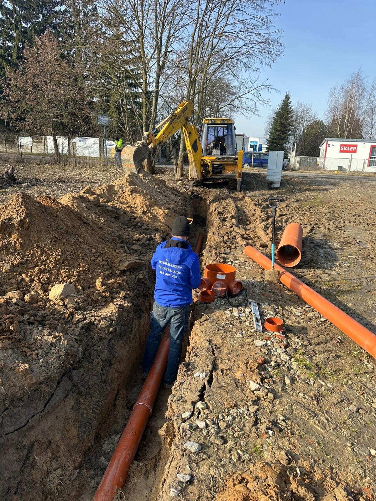 Montaż rur kanalizacyjnych w wykopie ziemnym z użyciem koparki. Widoczny pracownik w niebieskiej bluzie z napisem, pomarańczowe rury i akcesoria.