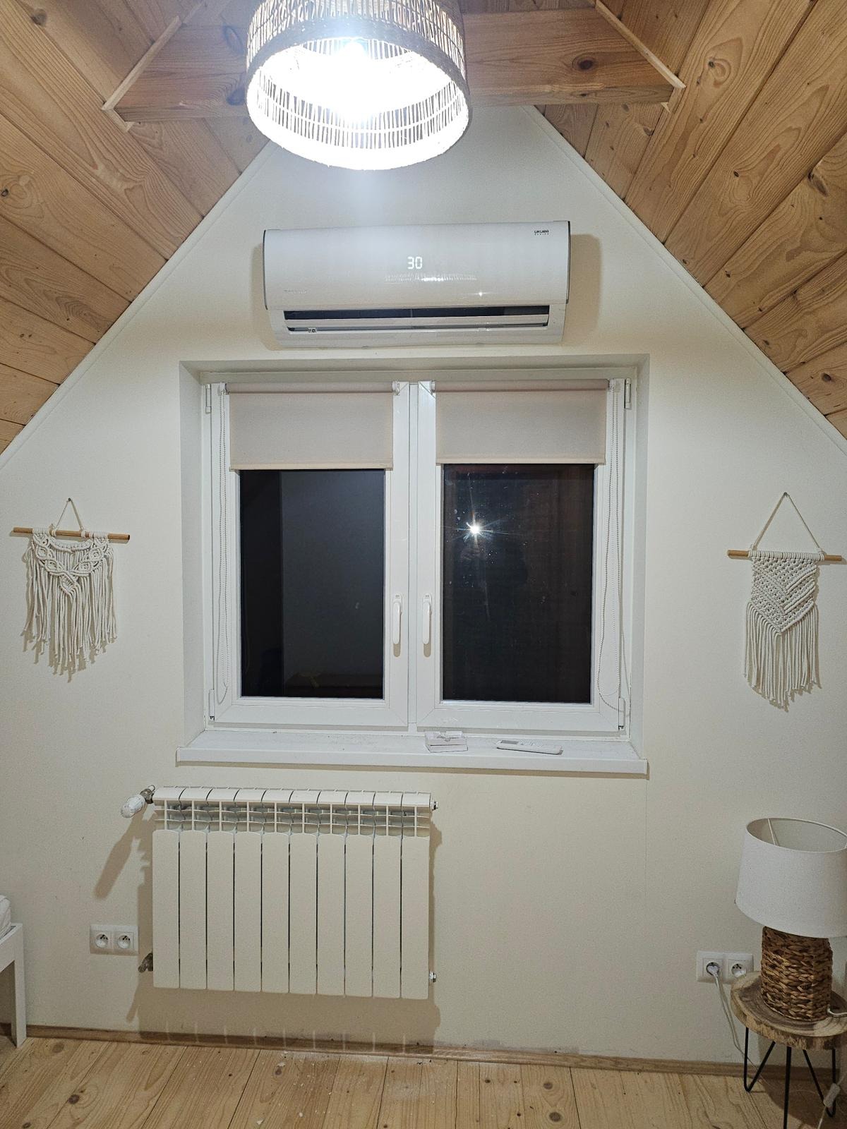 Biały grzejnik aluminiowy pod oknem na poddaszu z drewnianą boazerią, klimatyzator ścienny, lampa i makramy na ścianie.