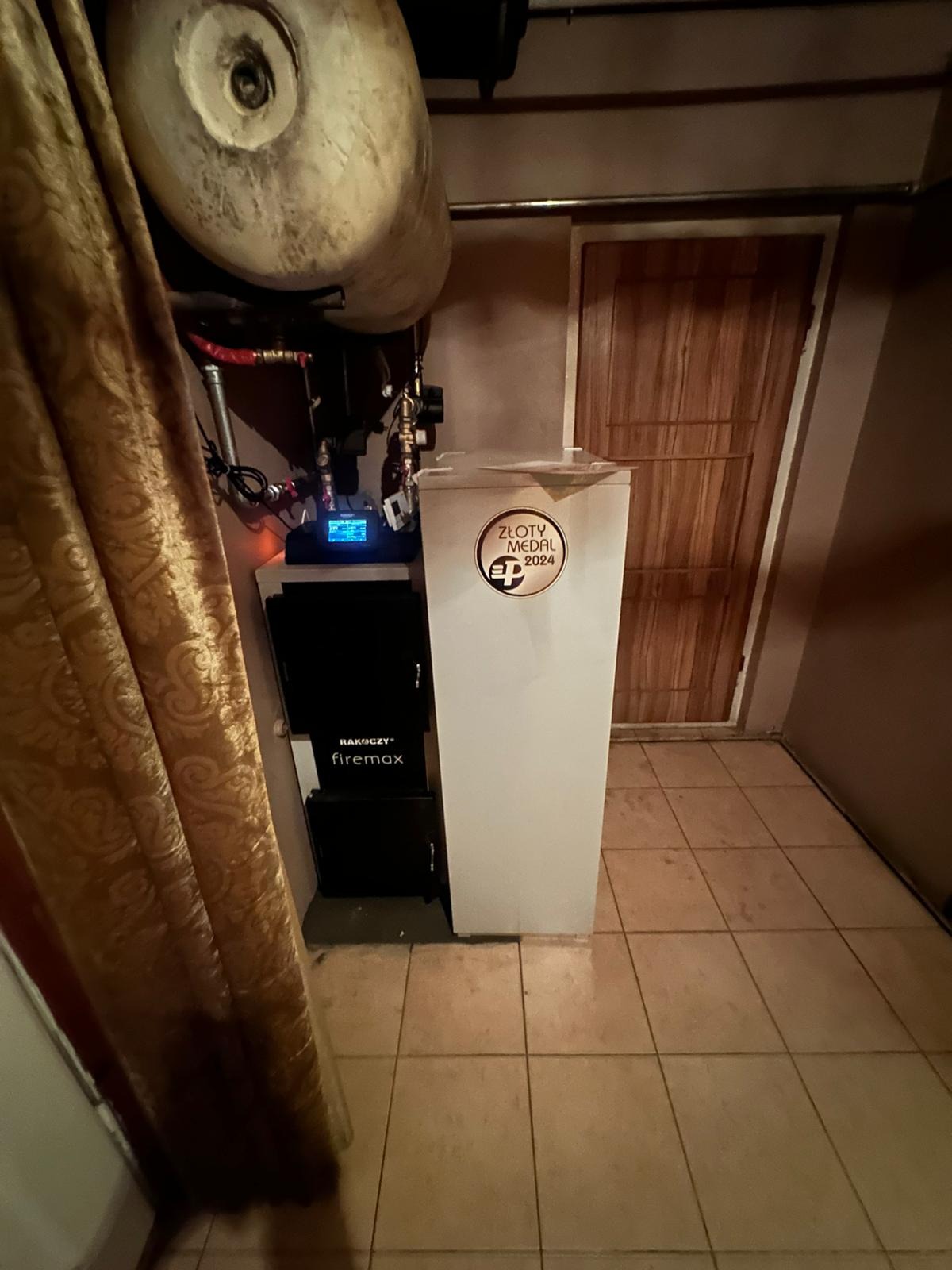 Zamontowany piec Rakoczy Firemax z oznaczeniem Złoty Medal 2024, widoczny zbiornik wyrównawczy i instalacja, w tle drzwi. Ujęcie z dołu, z zasłoną po lewej.