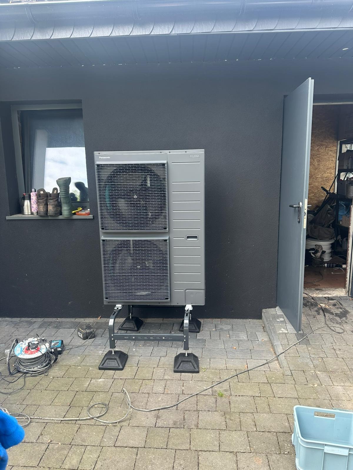 Szara pompa ciepła Panasonic Aquarea, zamontowana na specjalnej konstrukcji, stoi na kostce brukowej obok ściany budynku z oknem i otwartymi drzwiami.