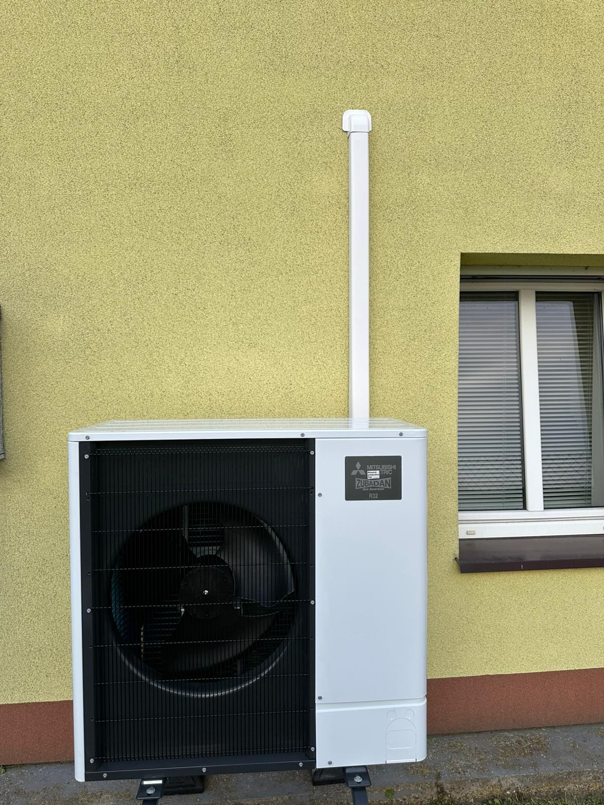 Biała pompa ciepła Mitsubishi Zubadan R32 zamontowana przy żółtej ścianie budynku z oknem. Widoczna biała rura odprowadzająca.