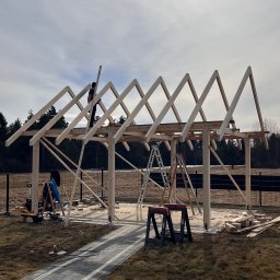 Józef Zubek Z BUD - Budowa drewnianej konstrukcji z widocznymi elementami więźby dachowej, drabiny i narzędzi. Prace na zewnątrz, w tle drzewa i ogrodzenie.