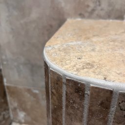 Józef Zubek Z BUD - Detale wykończenia: zaokrąglony narożnik obudowany mozaiką z trawertynu, wykończony białą fugą. Fragment ściany z płytkami w tle. Ujęcie z bliska.