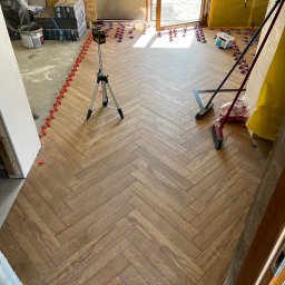 Józef Zubek Z BUD - Układanie parkietu w jodełkę z użyciem poziomicy laserowej i klipsów. Widoczne płytki, narzędzia glazurnicze i surowy stan wykończenia pomieszczenia.