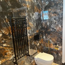 Józef Zubek Z BUD - Wykończona łazienka z płytkami imitującymi marmur w odcieniach czerni i brązu. Widoczny biały sedes, czarny grzejnik drabinkowy i złote akcesoria.