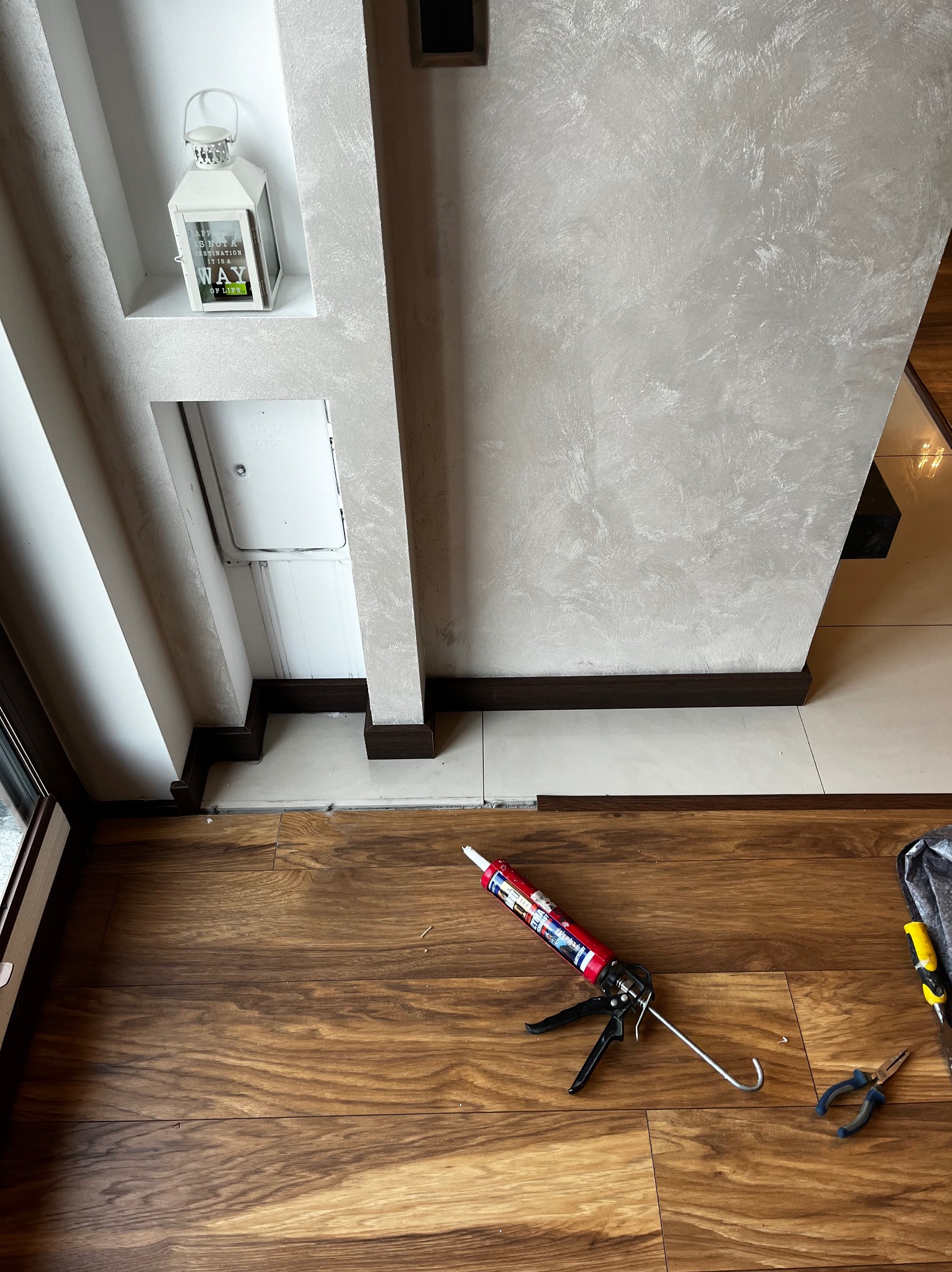 Montaż paneli podłogowych: pistolet do silikonu na drewnianej podłodze, przy ścianie z dekoracyjną wnęką i lampionem. Widoczne narzędzia i fragment podłogi z płytek.